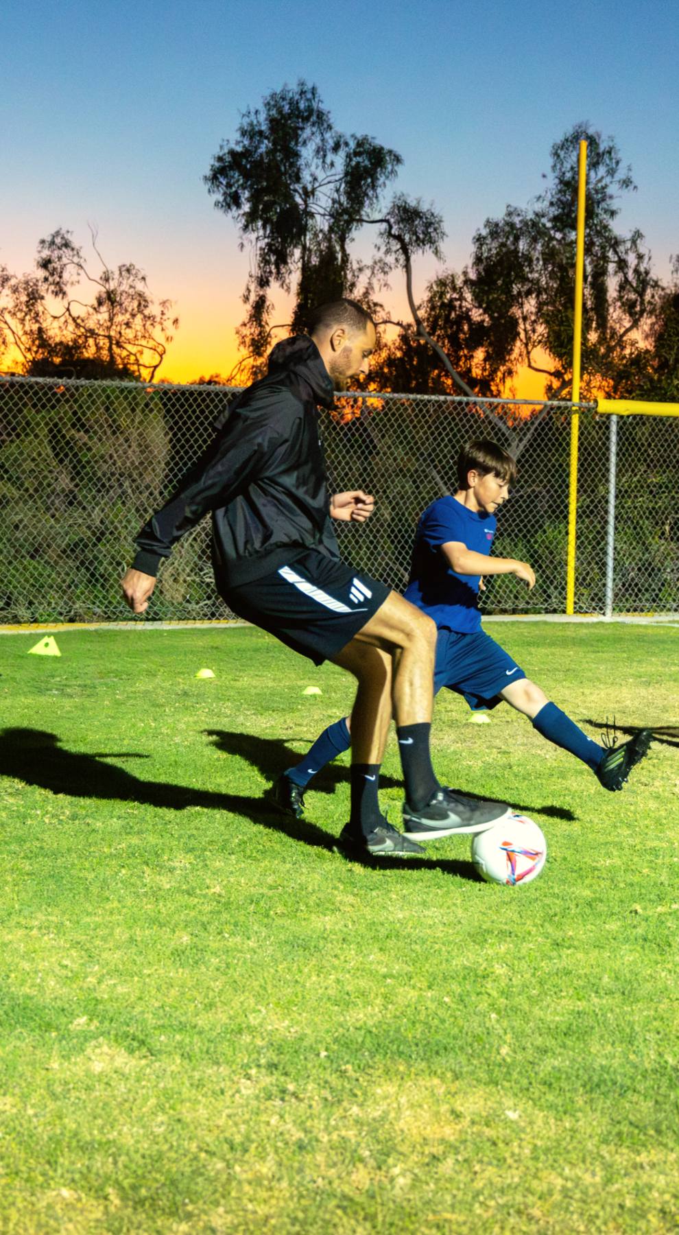 A young player strikes at the mini-goal under Coach Luka's pressure at dusk, the evaluation in action at The Soccer Lab, Carlsbad
